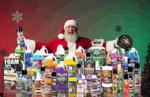 Santa Claus wearing a red suit and holding a Vapco brand hat and a can of Vapco Mongoose Coil Cleaner, standing in front of a festive display of Vapco products, celebrating the Holiday Cheer sweepstakes.